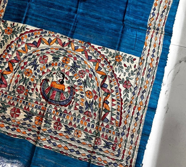 The Hand-Painted Madhubani Teal Blue Tussar Ghicha Silk Dupatta features intricate geometric and floral borders, with a central feminine medallion motif hand-painted and framed by decorative arches.